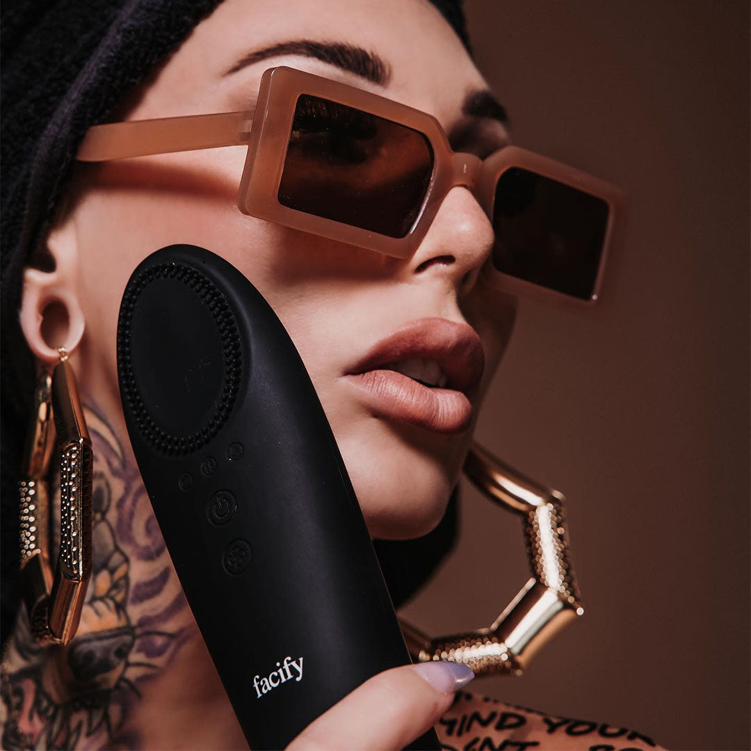 a woan wearing large sunglasses, big hoop earrings, and neck tattoos using the facify beauty wand on her face